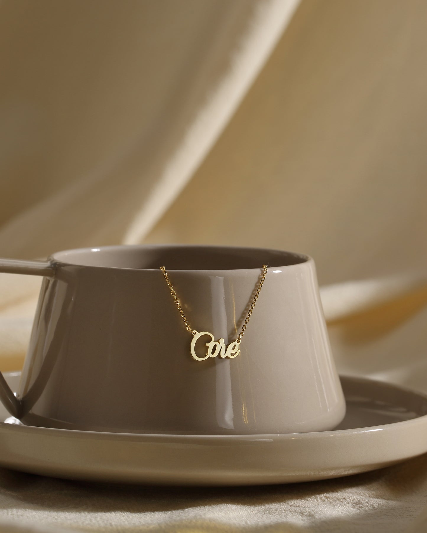 A personalized necklace with the inscription 'One' placed on a saucer and cup, suggesting a delicate and dainty design.