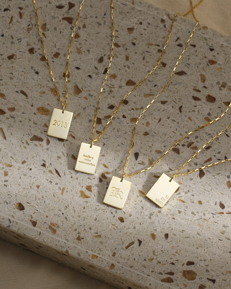 A collection of personalized necklaces with rectangular name plates on a gold-plated chain, displayed on a terrazzo surface.